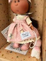 A close up of the handmade pink and white floral pinafore jumper dress over a cream muslin shirt and pink leatherette booties worn by a 9 inch vinyl doll named Little Mia Pink from Nines'd'Onil. The doll is situated in a star patterned cardboard box which is open at the front. 