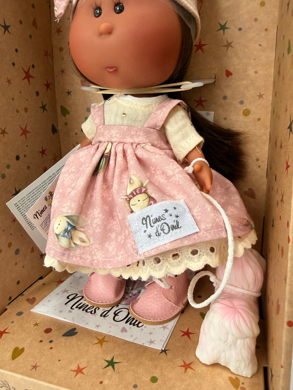 A close up of the handmade pink and white floral pinafore jumper dress over a cream muslin shirt and pink leatherette booties worn by a 9 inch vinyl doll named Little Mia Pink from Nines'd'Onil. The doll is situated in a star patterned cardboard box which is open at the front. 