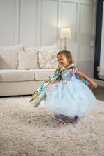 A young girl twirling in a light blue princess costume dress with a gold and blue cape, standing on a beige shag rug in a living room with a sofa and lamp in the background.