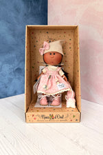 A vintage-style vinyl collectible doll named Little Mia Pink from Nines d'Onil. The doll has a light brown face with hand painted features and a little nose, long straight dark brown hair. She wears a pink and white floral pinafore jumper dress over a cream muslin shirt, pink leatherette shoes, and a beige knitted slouchy beanie with a pink bow matching her dress. She is accompanied by a small white dog with pink accents. She is in her own decorated display box.