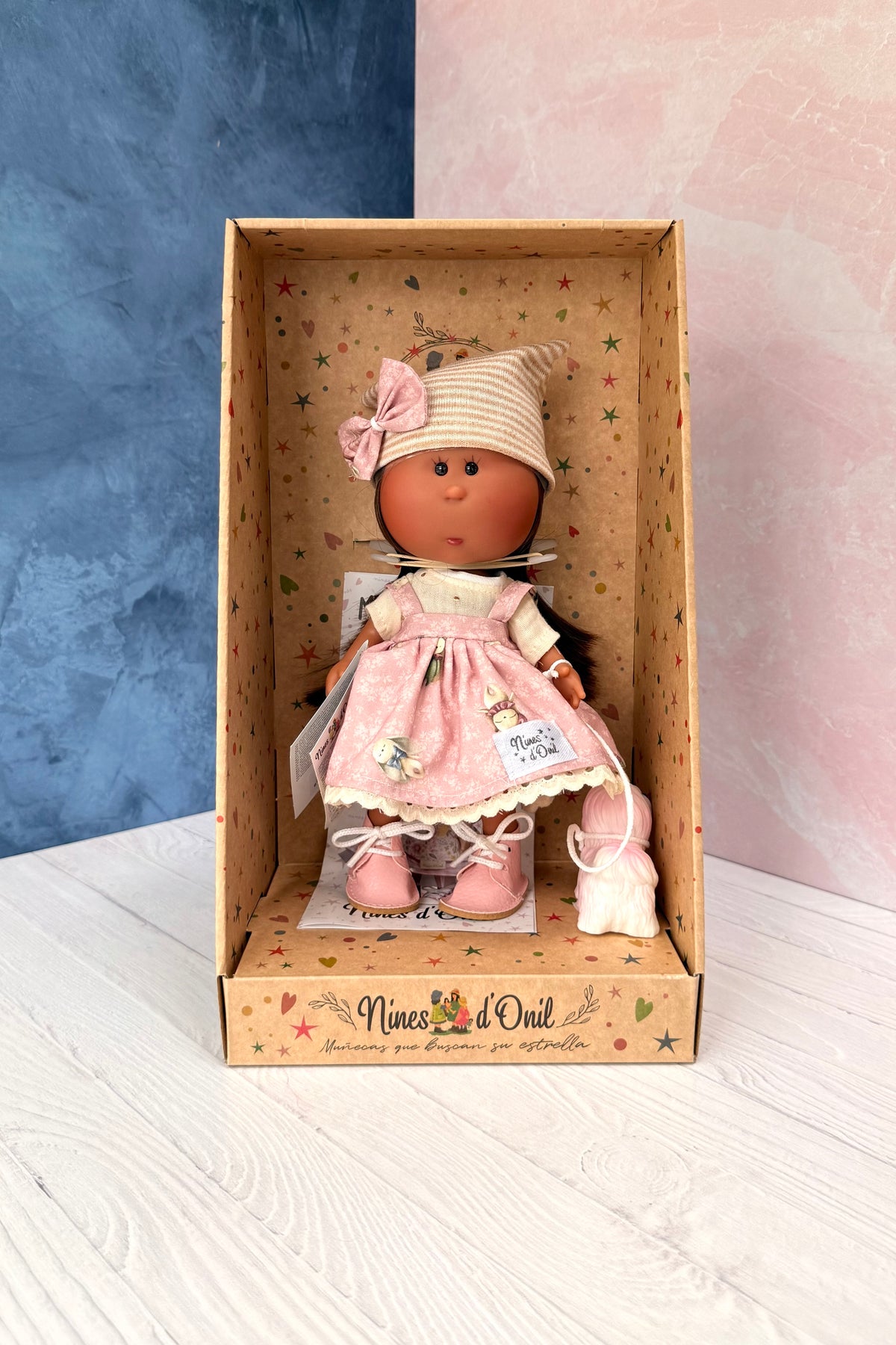 A vintage-style vinyl collectible doll named Little Mia Pink from Nines d'Onil. The doll has a light brown face with hand painted features and a little nose, long straight dark brown hair. She wears a pink and white floral pinafore jumper dress over a cream muslin shirt, pink leatherette shoes, and a beige knitted slouchy beanie with a pink bow matching her dress. She is accompanied by a small white dog with pink accents. She is in her own decorated display box.