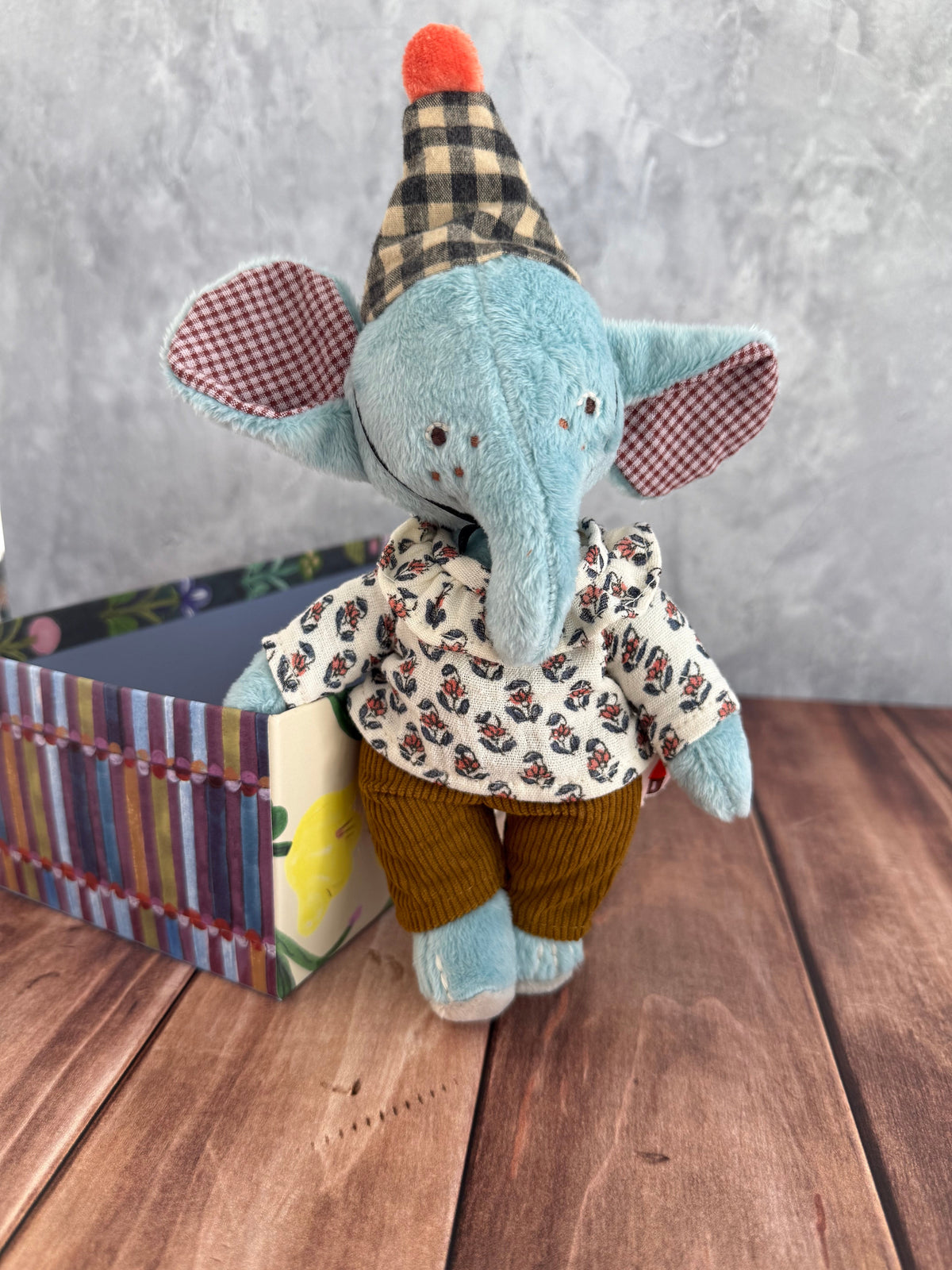 A plush, 10 inch elephant toy named Pablo the Elephant, part of Moulin Roty's Les Minouchkas line, is wearing a plaid fabric party hat with an orange pom at the top, an earth-toned floral patterned muslin shirt, and brown corduroy pants. His large ears are lined on the inside with playful red gingham linen.  He has black embroidered eyes and brown embroidered freckles. Pablo is posed next to his colorful, sturdy gift box, which houses him. They are posed on a wooden surface against a gray wall.