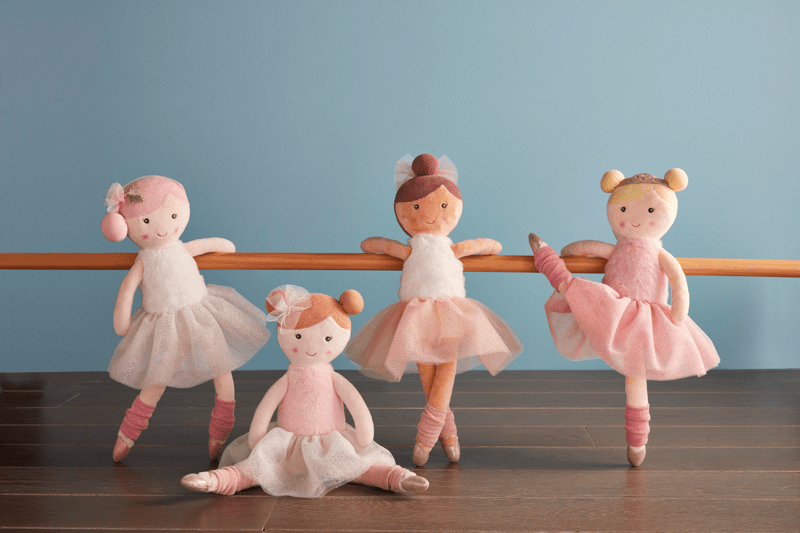 Four ballerina dolls in pink outfits standing on a wooden floor against a blue wall.