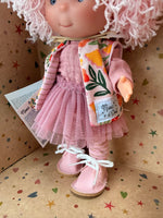 Close-up of the textured mauve shirt and a mauve layered crinoline "tutu" type skirt over mauve striped tights, pink leatherette shoes, and knit pink sleeveless sweater with a bright tropical print worn by a 9" doll named Little Pippa Pink Wool handmade by Nines d'Onil. Shown is the bottom part of her bountiful mop of pink loose curls. She stands in a star patterned display box which is open at the front.