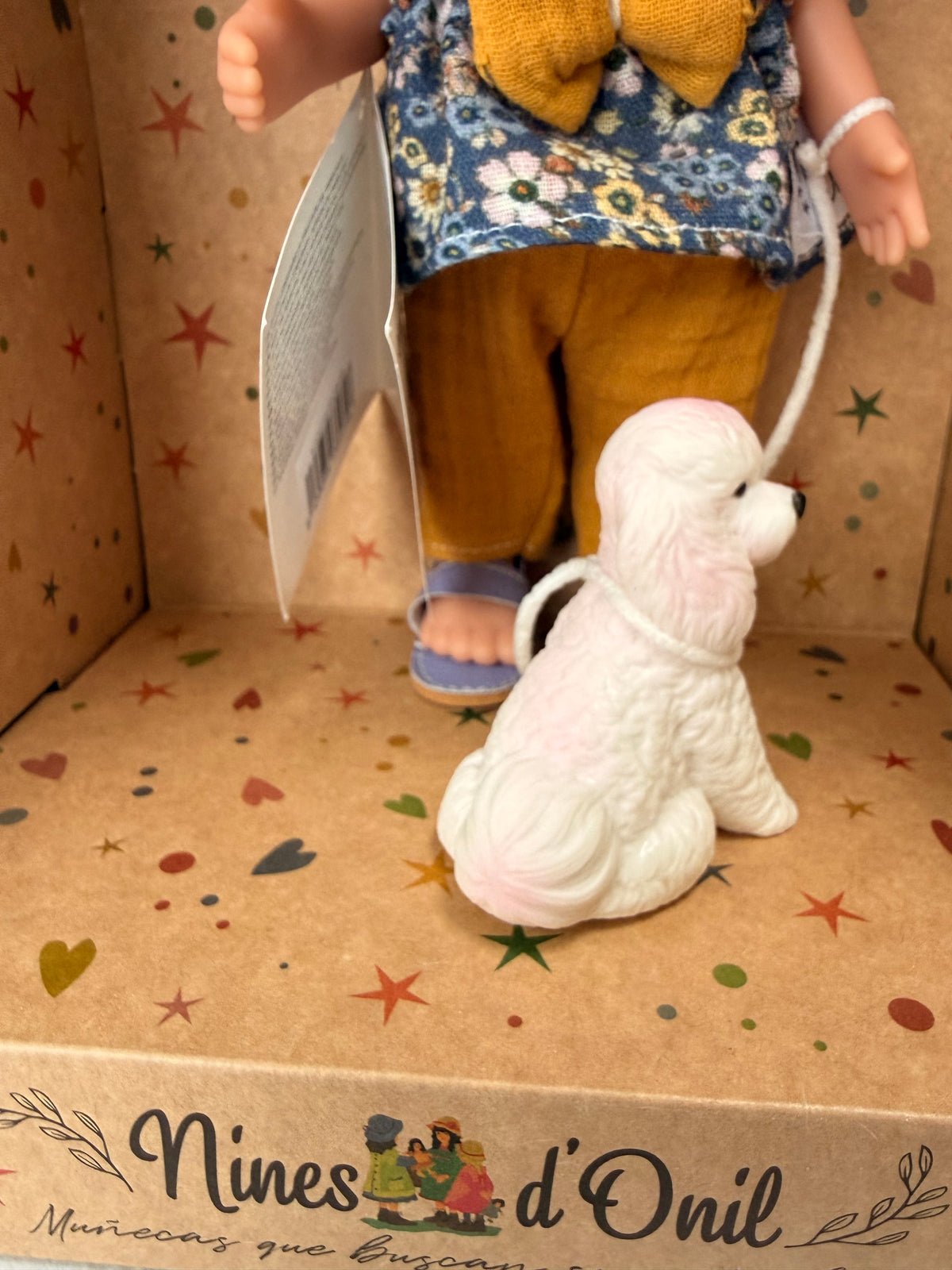 Close up of the back of a white and pink poodle type dog and the lower half of her handler, a 9" doll named Little Mia Walker, handmade in Spain by Nines d'Onil. Also pictured is her top, which is a blue floral sleeveless babydoll top with a large mustard yellow bow in the middle. She also wears textured mustard yellow pants and one lavender sandal can be seen. They are standing in a star patterned cardboard display box open in the front.