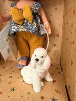 Close up of the front of a white and pink poodle type dog  and the lower half of her handler, a 9" doll named Little Mia Walker, handmade in Spain by Nines d'Onil. Also pictured is her top, which is a blue floral sleeveless babydoll top with a large mustard yellow bow in the middle. She also wears textured mustard yellow pants and one lavender sandal can be seen. They are standing in a star patterned cardboard display box open in the front.