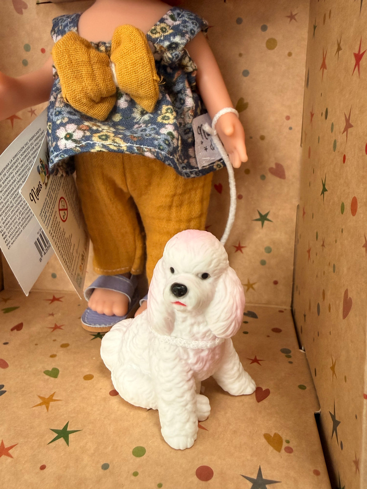 Close up of the front of a white and pink poodle type dog  and the lower half of her handler, a 9" doll named Little Mia Walker, handmade in Spain by Nines d'Onil. Also pictured is her top, which is a blue floral sleeveless babydoll top with a large mustard yellow bow in the middle. She also wears textured mustard yellow pants and one lavender sandal can be seen. They are standing in a star patterned cardboard display box open in the front.