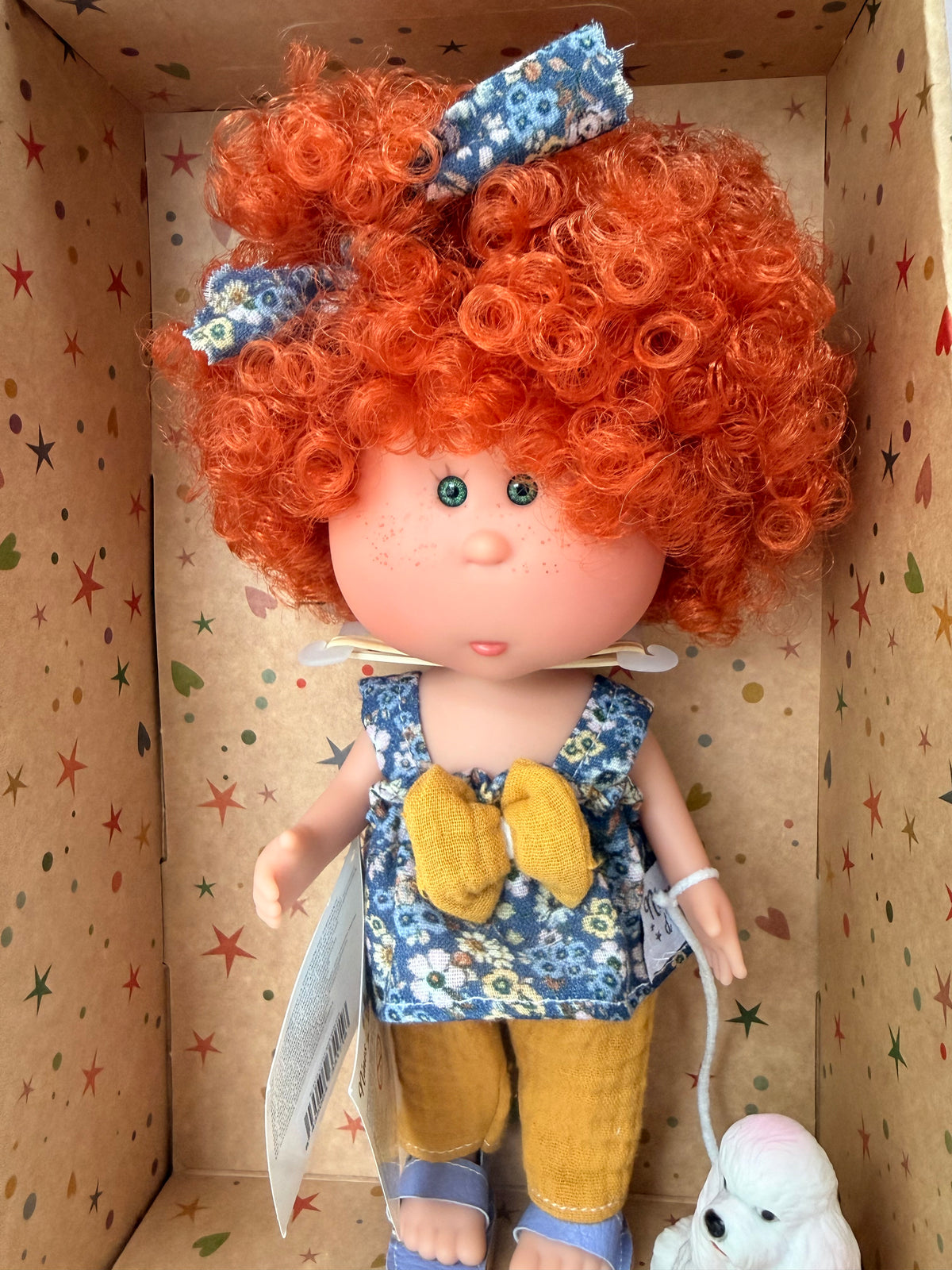Close up of a 9" doll named Little Mia Walker, handmade in Spain by Nines d'Onil. This closeup shows tiny freckles on Little Mia's hand painted face and bright green eyes. She wears a blue floral sleeveless babydoll top with a large mustard yellow bow in the middle. She also wears textured mustard yellow pants and lavender sandals. They are standing in a star patterned cardboard display box open in the front.