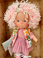 Close-up of a 9" doll named Little Pippa Pink Wool handmade by Nines d'Onil. She has a bountiful mop of pink loose curls on her head, which features a silver barrette in the shape of a star. She wears a textured mauve shirt and a mauve layered crinoline "tutu" type skirt over mauve striped tights, and pink leatherette shoes. She wears a knit pink sleeveless sweater with a bright tropical print.  She stands in a star patterned display box which is open at the front.