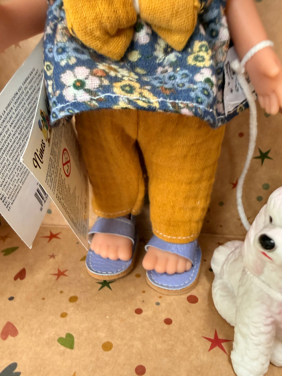 Close up of the mustard yellow pants and lavender sandals worn by a 9" doll named Little Mia Walker, handmade in Spain by Nines d'Onil. Also pictured is the bottom portion of her top, which is a blue sleeveless babydoll top with a large mustard yellow bow in the middle. A portion of the white and pink doll that accompanies her is also seen in this close-up. They are standing in a star patterned cardboard display box open in the front.
