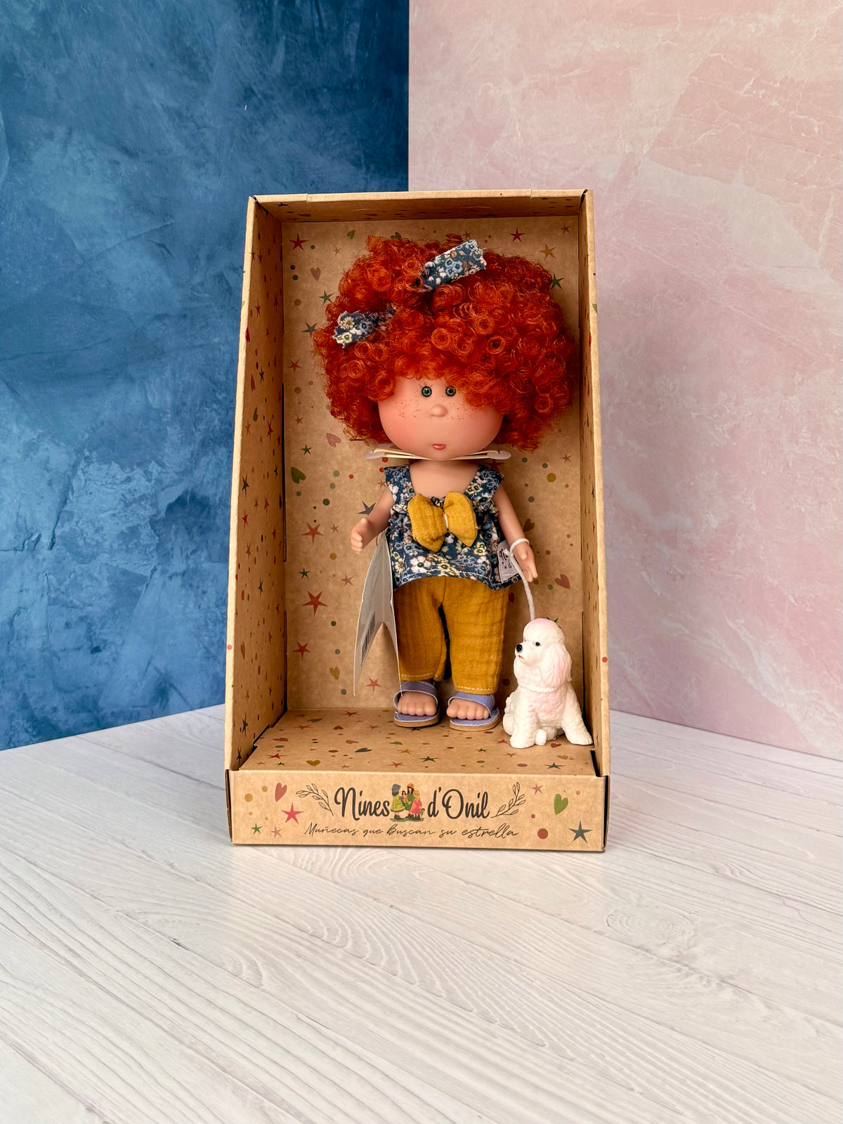 9" doll named Little Mia Walker, handmade in Spain by Nines d'Onil. She has a halo of short, red curly hair with a headband that matches her top, which is a blue sleeveless babydoll top with a large mustard yellow bow in the middle, along with textured mustard yellow pants and lavender sandals.  She is accompanied by a small white and pink dog. They are standing in a star patterned cardboard display box open in the front.