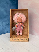 A 9" doll named Little Pippa Pink Wool handmade by Nines d'Onil. She has a bountiful mop of pink loose curls on her head, which features a silver barrette in the shape of a star. She wears a textured mauve shirt and a mauve layered crinoline "tutu" type skirt over mauve striped tights, and pink leatherette shoes. She wears a knit pink sleeveless sweater with a bright tropical print.  She stands in a star patterned display box which is open at the front.
