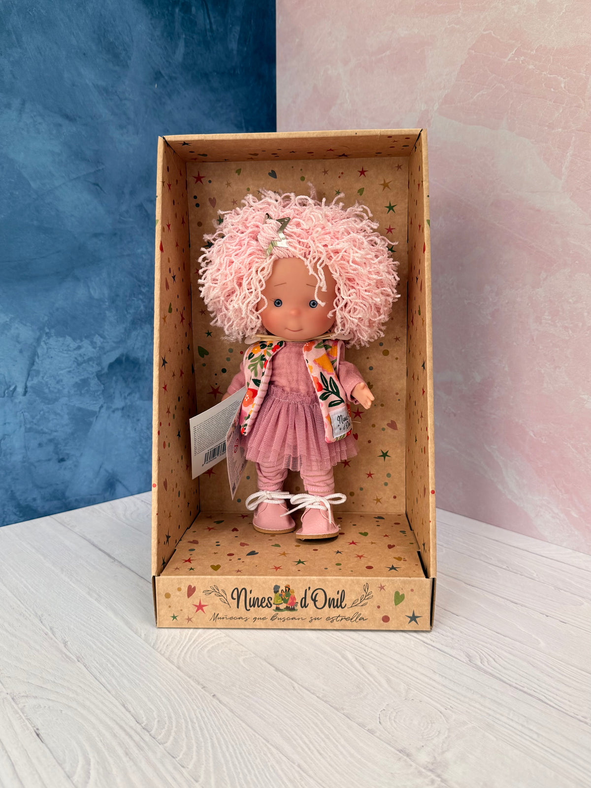 A 9" doll named Little Pippa Pink Wool handmade by Nines d'Onil. She has a bountiful mop of pink loose curls on her head, which features a silver barrette in the shape of a star. She wears a textured mauve shirt and a mauve layered crinoline "tutu" type skirt over mauve striped tights, and pink leatherette shoes. She wears a knit pink sleeveless sweater with a bright tropical print.  She stands in a star patterned display box which is open at the front.