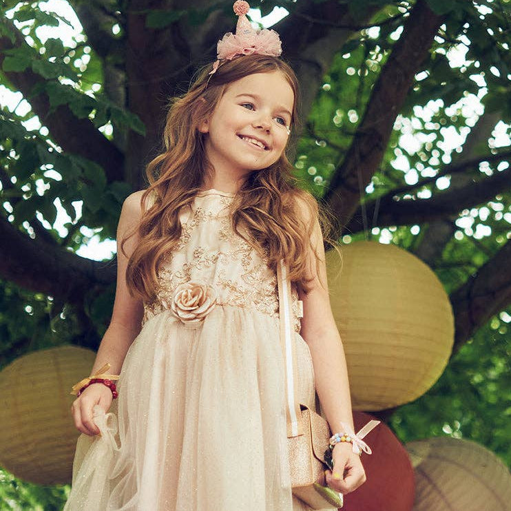A young girl with long wavy brown hair stands under a tree wearing a pale blush pink tulle dress with floral embroidery and a matching headpiece. She holds a small white handbag and stands beside hanging paper lanterns.