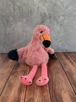 A pink plush flamingo toy from Warmies with a black and orange beak, sitting on a dark wood floor against a textured gray wall.