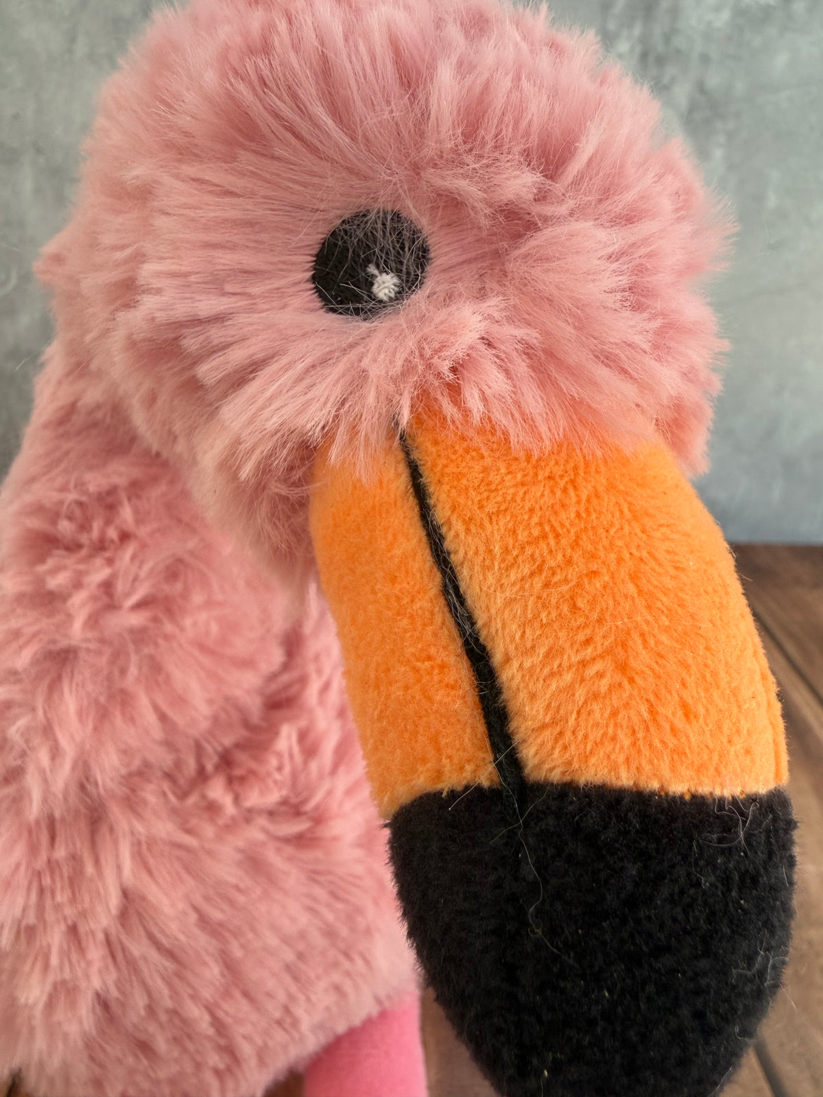 A close-up of a plush flamingo toy by Warmies with fluffy pink feathers, a bright orange beak, and a black nose and eye detail, resting on a wooden surface against a gray wall.