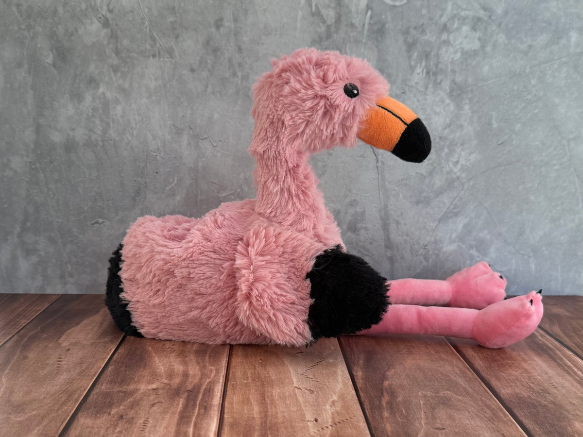 A pink plush flamingo toy from Warmies with black accents on its legs and feet, and an orange and black beak, sitting on a wooden surface against a textured gray wall.