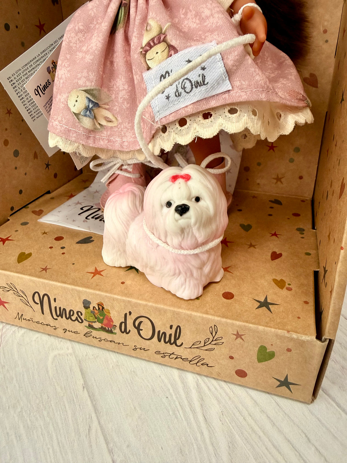 A close up of a small white and pink long haired vinyl dog that accompanies collectible Little Mia Pink, a 9 inch doll. The dog is boxed along with the doll in a star patterned decorated cardboard display box that is open at the front