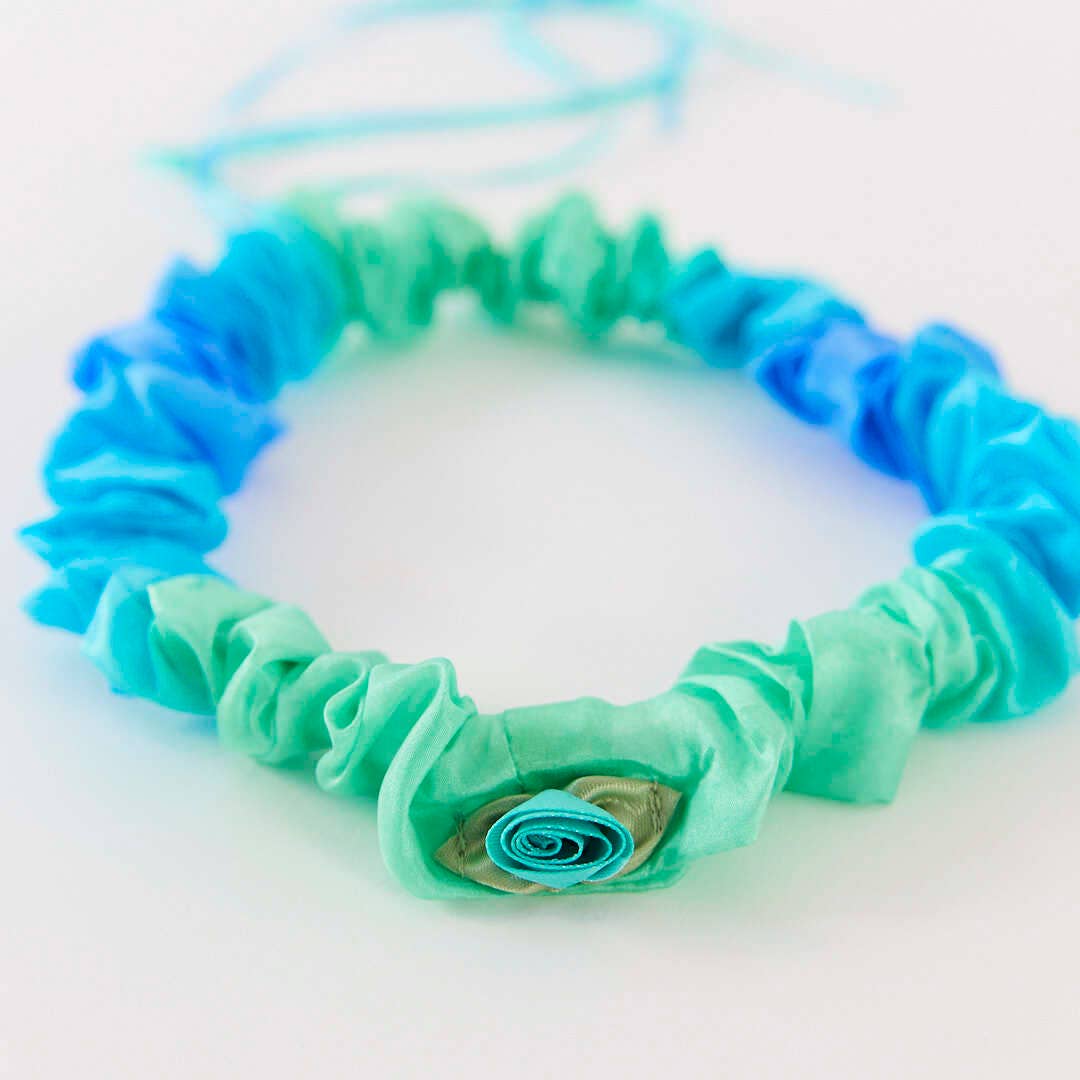 A handmade fabric hair bow or hair tie made from ruffled layers of mint green and blue ribbon, featuring a small rolled fabric flower accent at the center, shown on a plain white background.