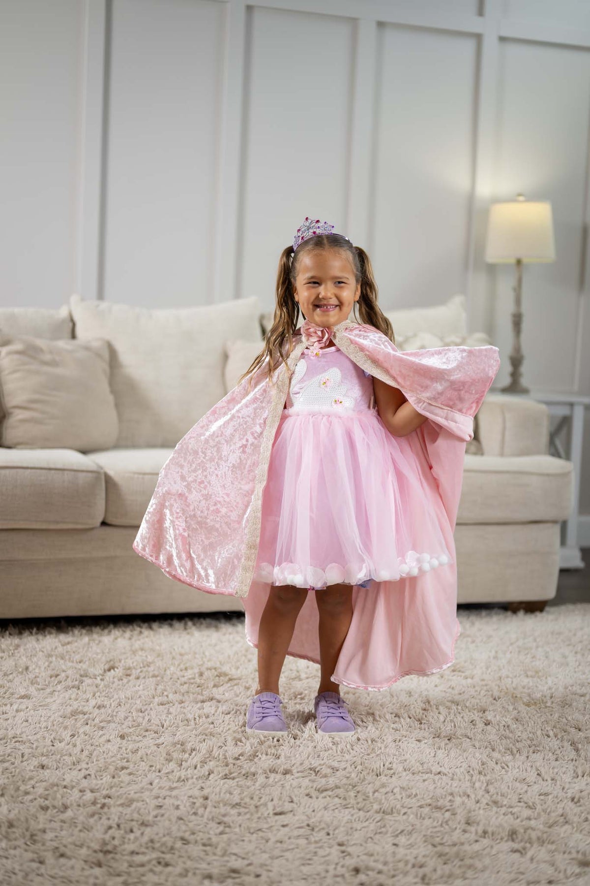 A young girl stands in a living room wearing a pink princess costume with a matching cape and tiara. The costume features a white cartoon character print on the chest and a layered pink tulle skirt.